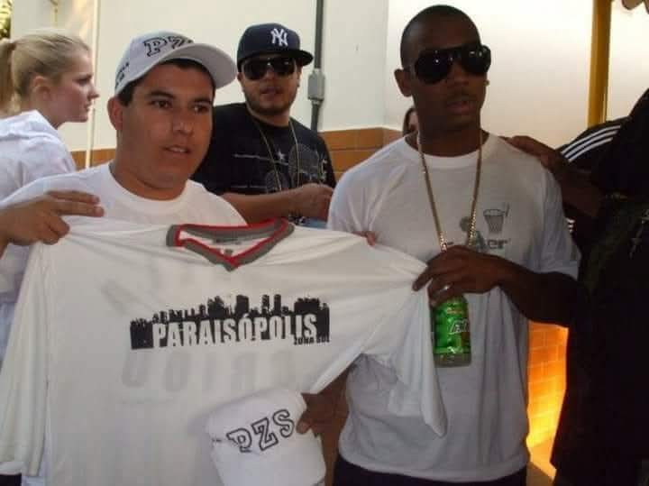 Guga ao lado de Ja Rule, rapper americano segurando a camisa da marca “A Favela me Criou”